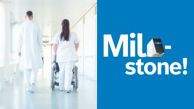 A hospital corridor, in which a man and a woman in white work clothes can be seen from behind. The woman is pushing a wheelchair.