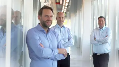 A photo of three men standing with their arms crossed in a long, brightly lit hallway. They are wearing light-blue shirts and dark trousers and smile relaxed at the camera.